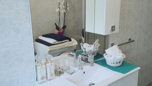 Ein Badezimmer mit einem weißen Waschbecken, Spiegel und Ablagekorpus. Auf der Ablage liegen Toilettenartikel, ein Handtuch und eine Vase mit Blumen.