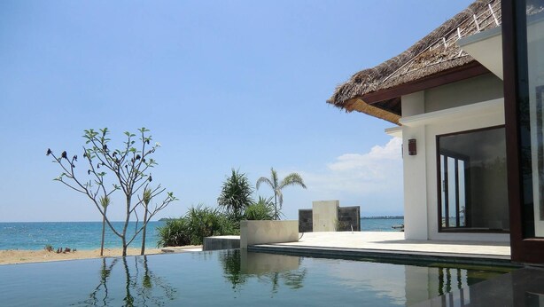Una piscina moderna con vista despejada al océano, rodeada de plantas tropicales y un edificio con techo de paja.