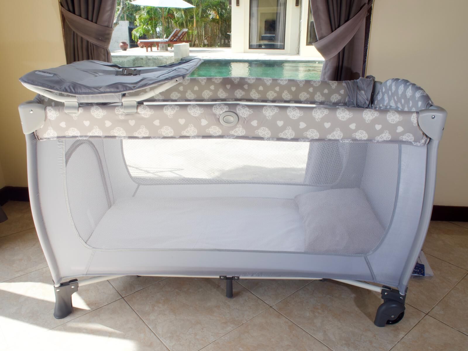 Un parc pour bébé portable, doté d’une paroi en tissu à mailles, d’un dessus rembourré et de roulettes, installé à l’intérieur et offrant une vue sur une piscine extérieure.