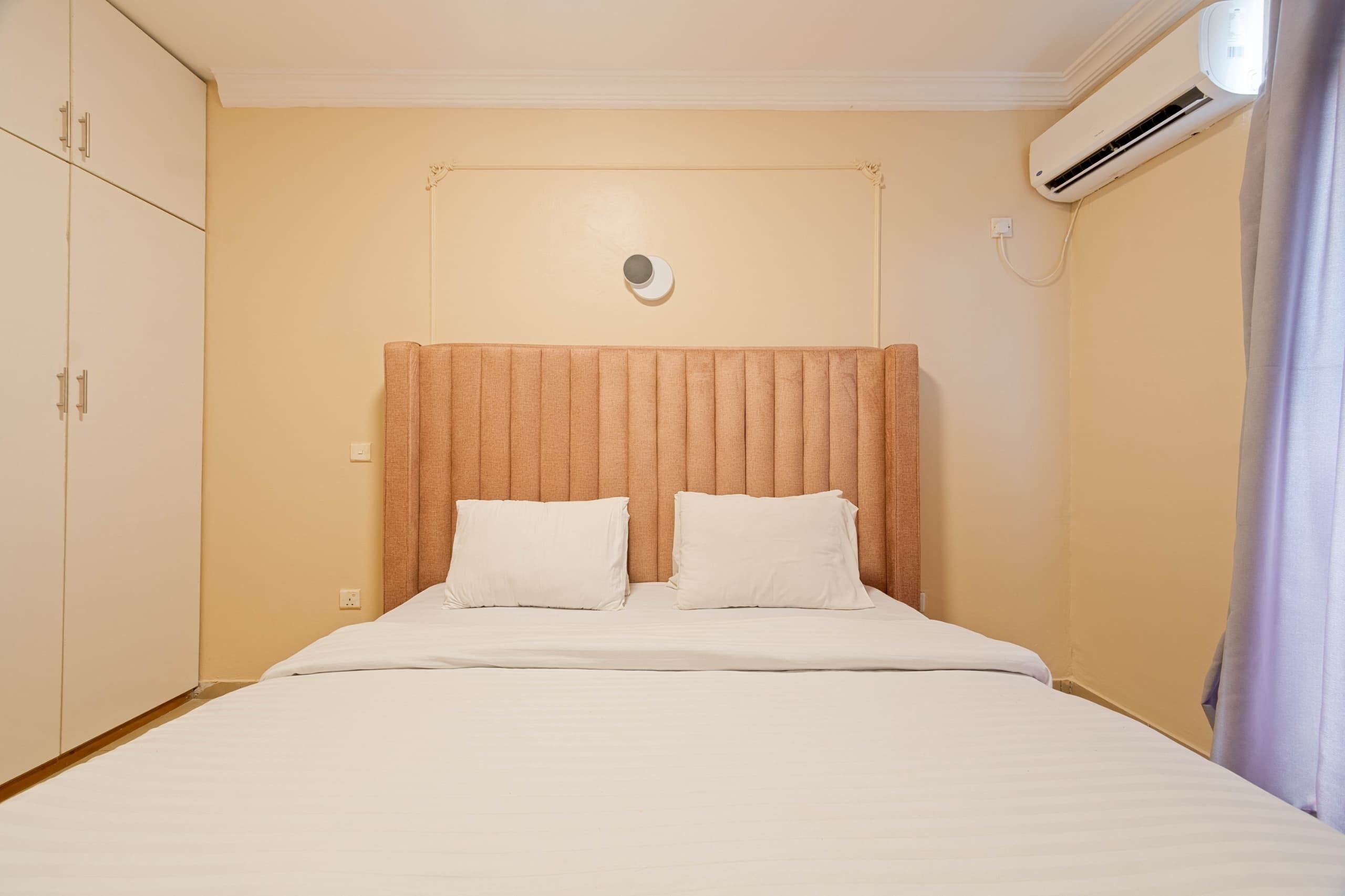 A hotel room with a bed, a headboard, a wardrobe, and an air conditioning unit.