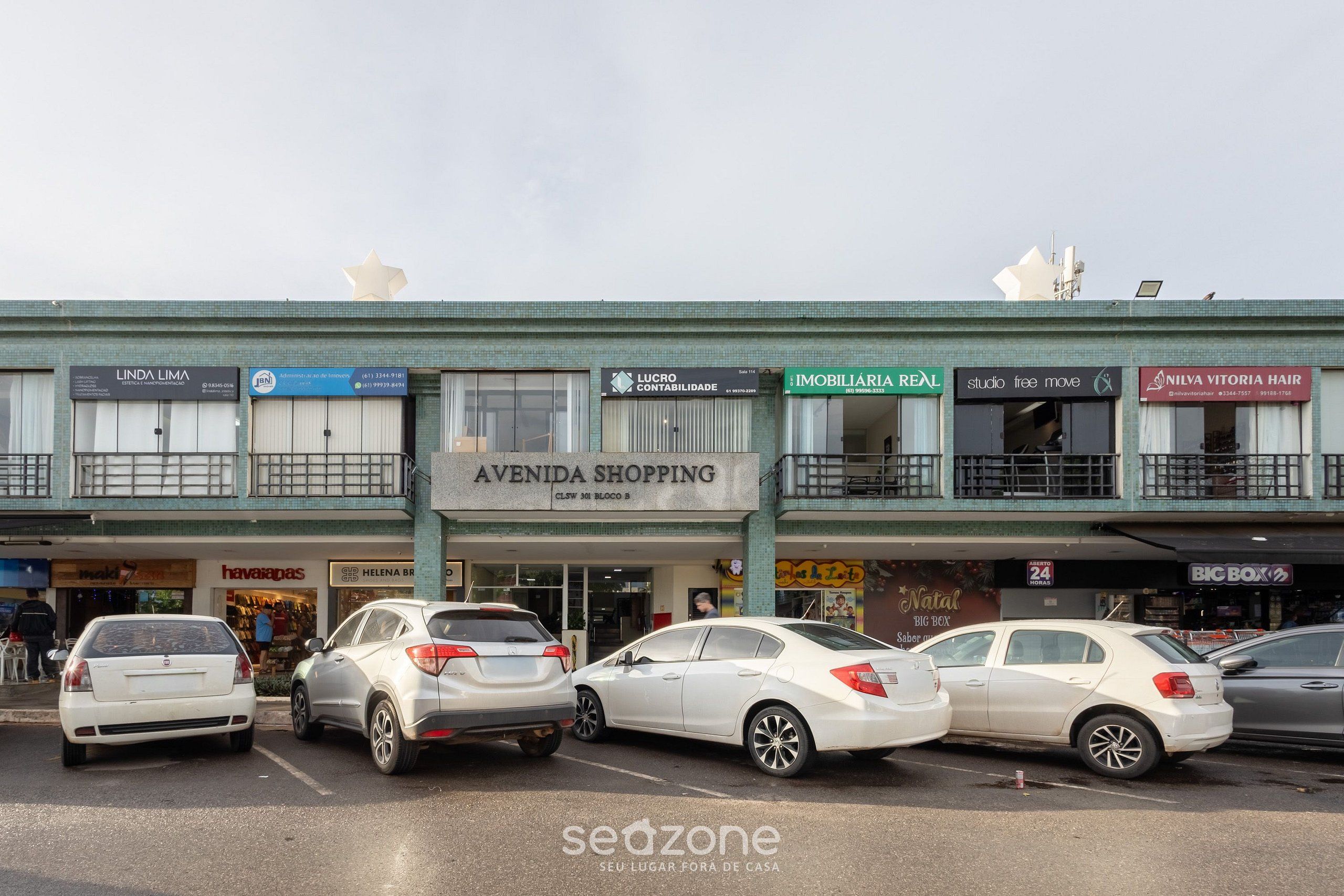 A street view with multiple storefronts, including 'Avenida Shopping' and 'Imobiliaria Real'. Several cars are parked along the street.