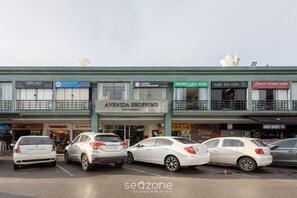 A street view with multiple storefronts, including 'Avenida Shopping' and 'Imobiliaria Real'. Several cars are parked along the street. - Modern apt in Sudoeste w/ A/C & Wi-fi Cls0101 (Sudoeste e Octogonal)
