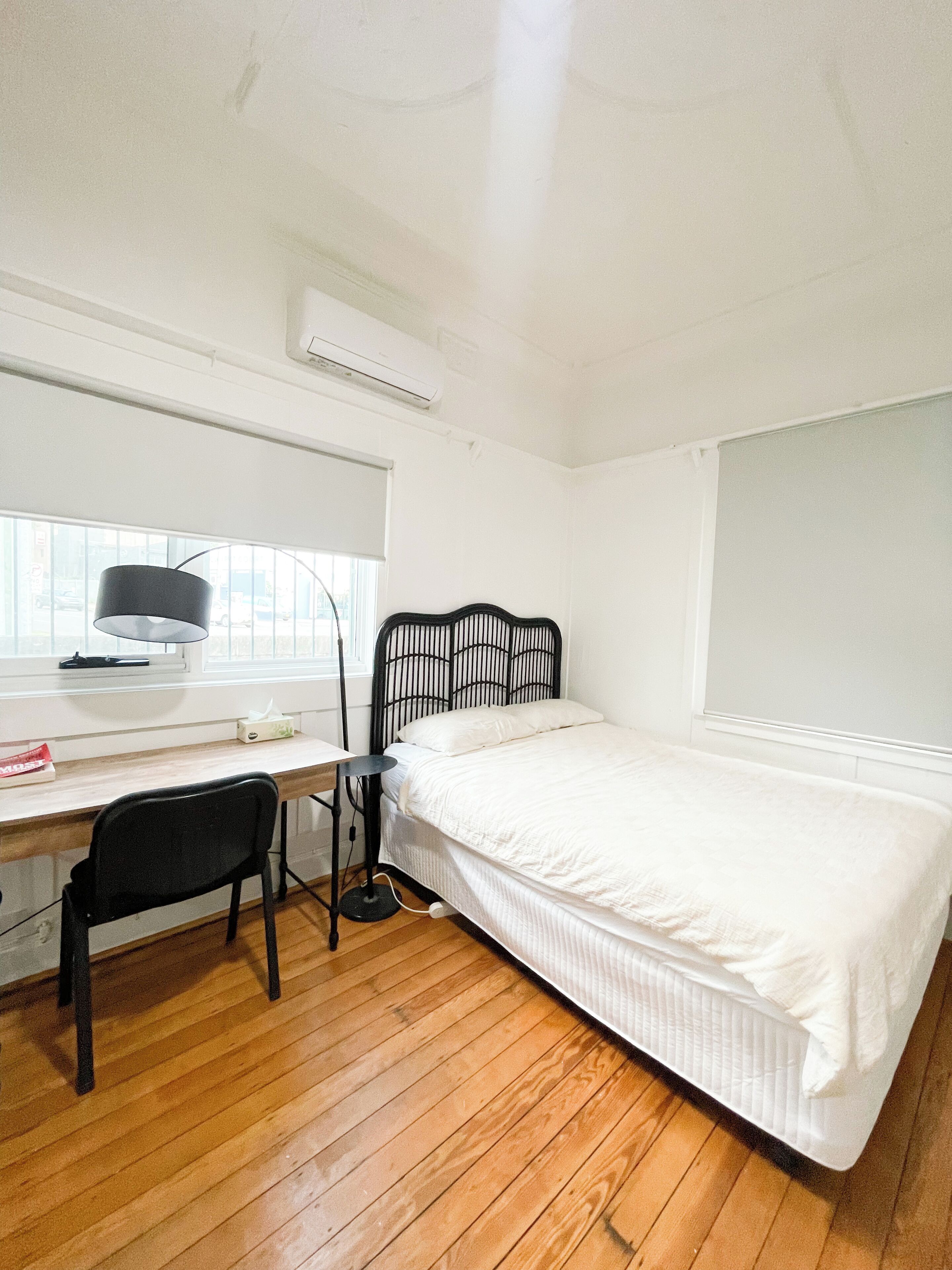 A bedroom with a wooden floor, a bed with white bedding, a black metal headboard, a desk with a lamp, and a chair.