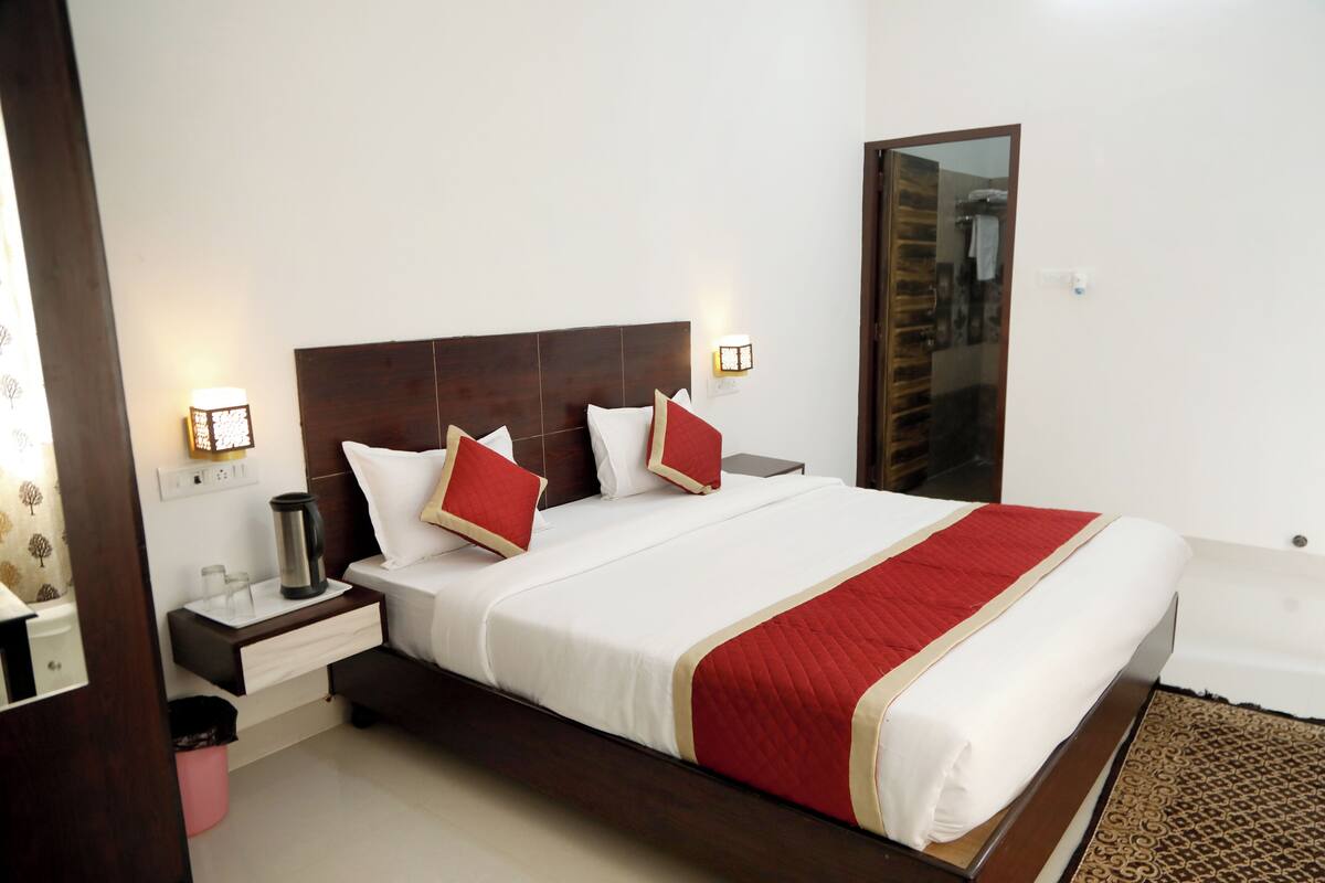 A hotel room with a large bed, bedside tables, a nightstand, a red and white bedspread, and a wooden headboard.