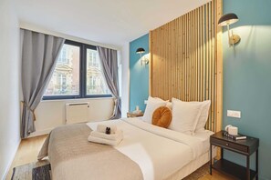 Ein Hotelzimmer mit Bett, Nachttisch, Lampe, einem Fenster mit Vorhängen und Blick auf Gebäude.
