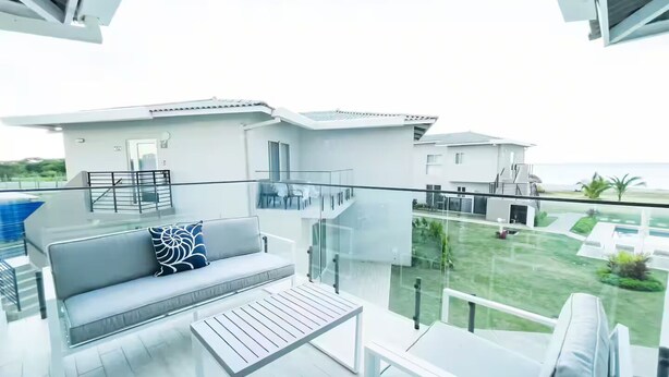 Un balcón moderno al aire libre con un sofá blanco, una mesa de centro de cristal y vistas a una zona residencial con piscina y vegetación.
