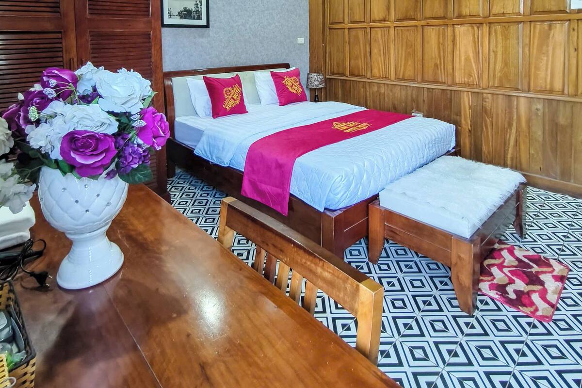 Ein Hotelzimmer mit Holzboden, einem Bett mit rotem und weißem Überwurf, einer hölzernen Bank, einem Tisch mit Blumenvase und einem gemusterten Boden.
