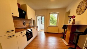 Een keuken met houten vloer, witte kastjes, een klok en een raam met uitzicht op groen.