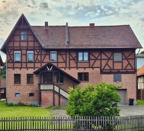 Ein traditionelles Fachwerkhäuschen mit einem steilen Satteldach, einem zentralen Eingangsbereich mit Treppe und einer kleinen Veranda.