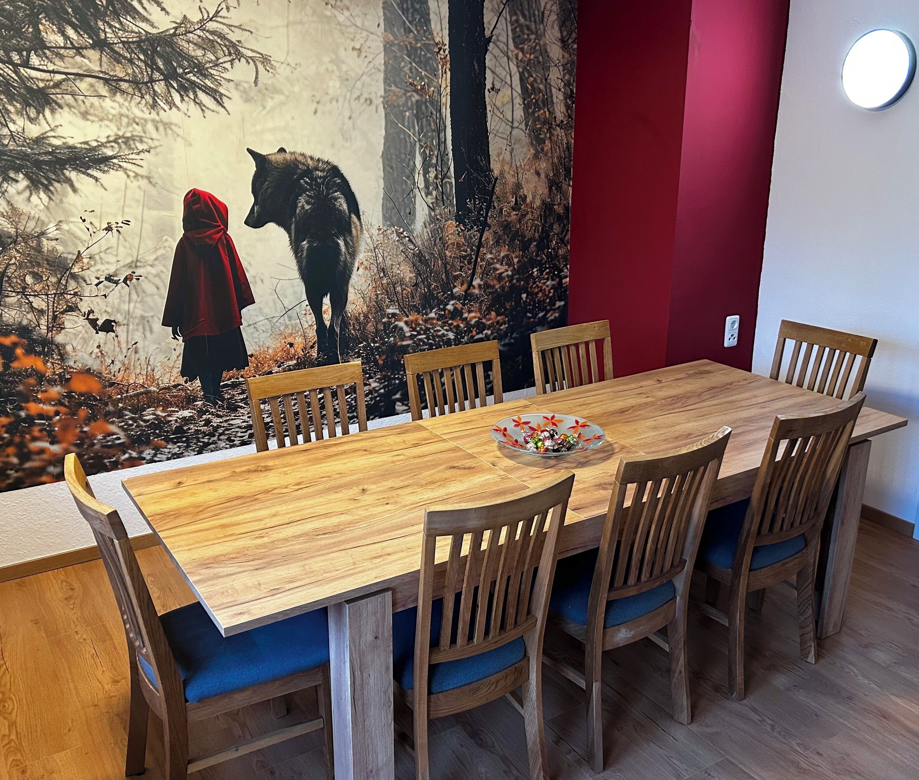 A dining area with a wooden table and chairs, a red accent wall, and a mural of a forest scene.