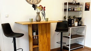 Een kleine keuken met een houten tafel, twee zwarte barstoelen, een grote wandklok en een wit schap met keukengerei.