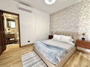 A bedroom with a bed, bedside tables, a nightstand, a patterned wallpaper, and a wooden floor. - 2-bedroom apartment in Downtown Punta Cana with private pool , AC and gym access (Punta Cana)
