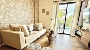 Ein Wohnzimmer mit einem beigen Sofa, gemusterten Poufs, einem kleinen runden Tisch und einem Fernseher an der Wand.