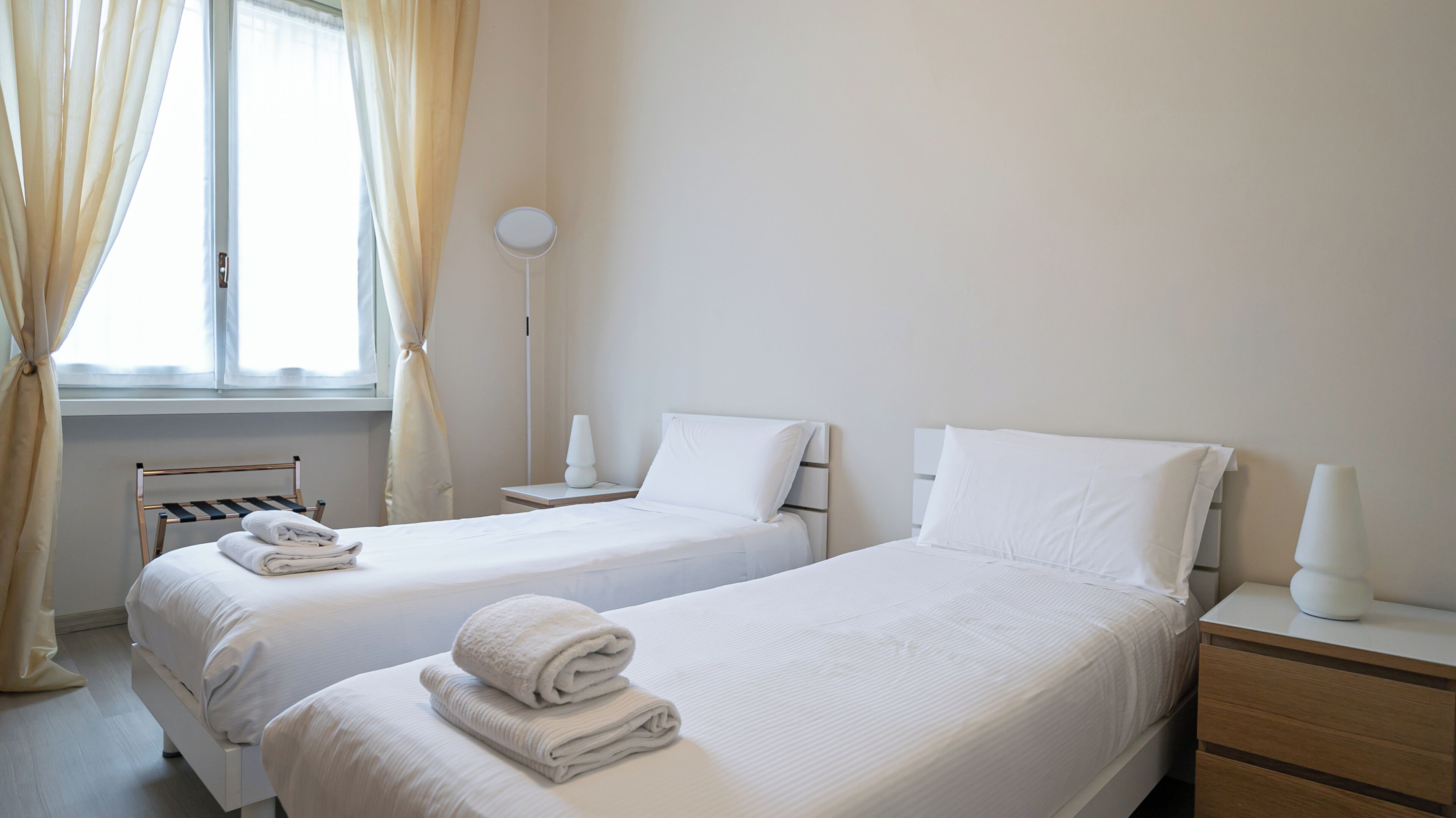 Et hotelværelse med to senge, et natbord, en lampe og et vindue med gardiner.