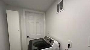 Ein Waschraum mit einer Waschmaschine und einem Trockner, einer Tür und einem Lüftungsschacht an der Wand.