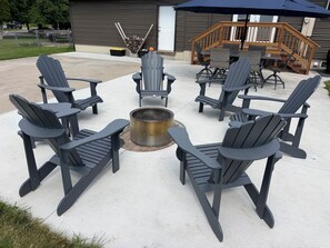 A patio with Adirondack chairs arranged around a fire pit, a wooden deck, and a garage door in the background. - 5-bedrooms, Tons of Outdoor Space, Central Location! (Kalispell)