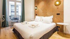 A hotel room with a large bed, a wooden accent wall, a small round table, and a view of buildings through a window with curtains. - HOTEL BOETIE - CHAMPS ELYSEES  (Paris)
