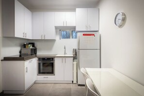Een kleine keuken met witte kastjes, een magnetron, een oven, een koelkast en een klein raam.