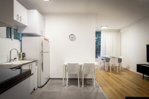 A modern kitchen with white cabinets, a refrigerator, a sink, and a coffee maker. There is a dining area with a table and chairs, and a TV on the right. - Cozy 1-Bedroom Retreat in Tel Aviv by SeaNRent (Tel Aviv-Yafo)