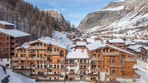 Ein verschneites Bergdorf mit Holzhäusern, darunter mehrstöckige Gebäude mit Balkonen und einem zentralen Gebäude mit schneebedecktem Dach.