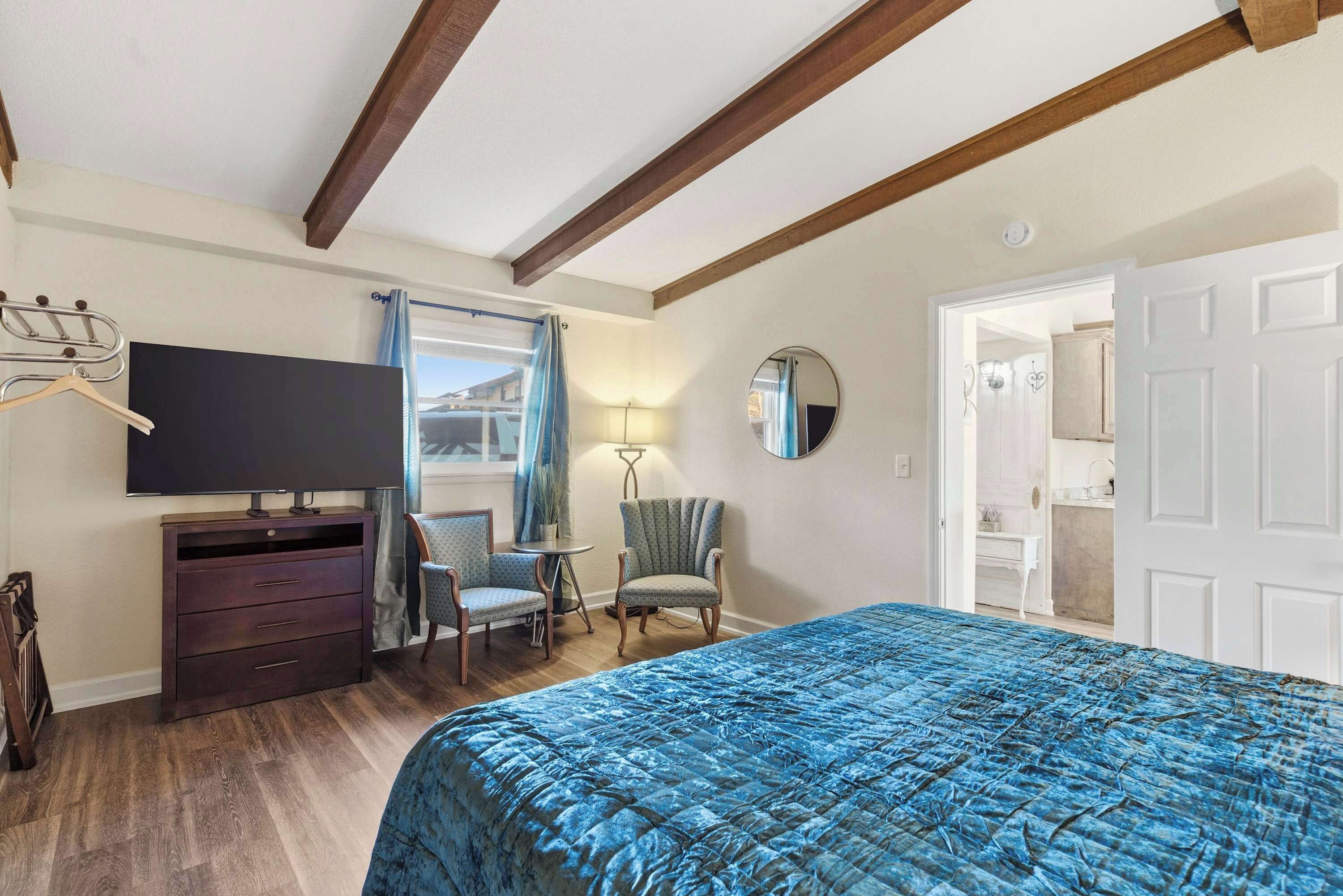 A bedroom with a bed, a TV, a dresser, two chairs, a mirror, and a door leading to a bathroom.