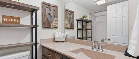 A bathroom with a beige countertop, a white sink, a mirror, and wooden shelves with decorative items.