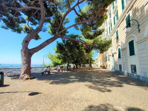 Eine von Bäumen gesäumte Promenade mit Bänken, freier Meeresblick und einem Gebäude im klassizistischen Stil.