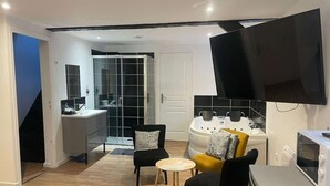 A modern living room with a sofa, chairs, a small table, a TV, and a kitchen area with a microwave and a white appliance. - Le Nid Cosy 4, Balnéo 2 lits wifi (Tourcoing)