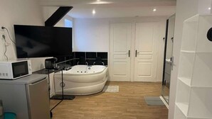 Een moderne hotelkamer met een bed, een klein keukentje, een ruime badkuip en een aan de muur bevestigde televisie.