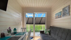 Interior acolhedor de uma cabana com um sofá, uma mesa de jantar e vista para uma área gramada através de uma porta de correr.