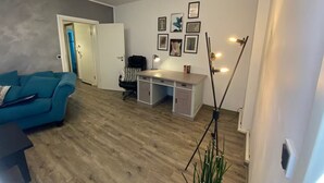 Un salon moderne avec un canapé bleu, un bureau et une chaise. Des tableaux encadrés ornent le mur, et une grande lampe sur pied se trouve dans un coin.