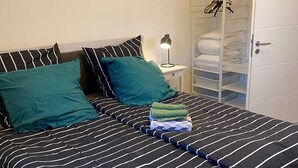 Un lit bien fait, avec une literie à rayures, une lampe de chevet et une armoire où sont suspendus des vêtements.