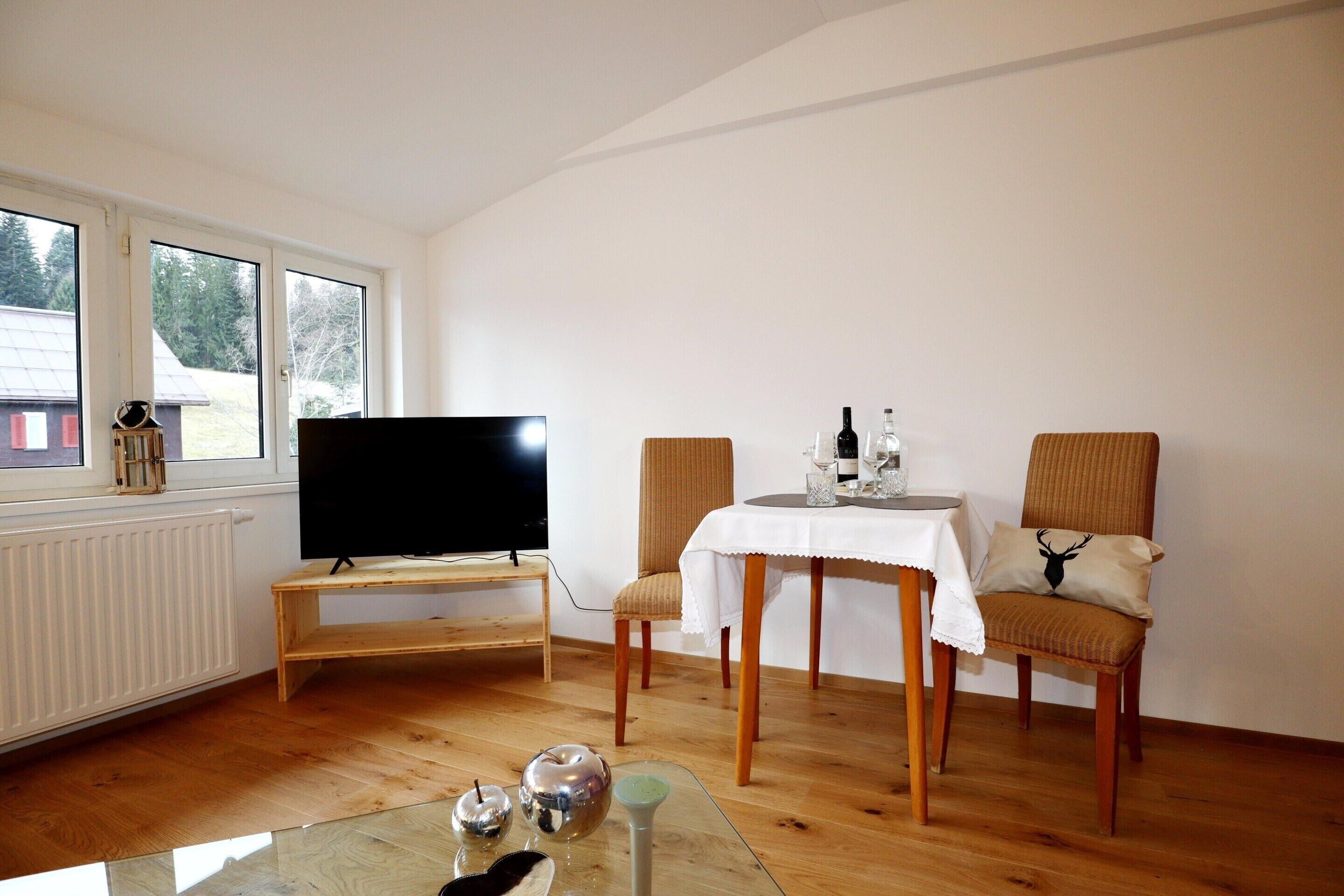 A room with a television on a wooden stand, a table with a white tablecloth, two chairs, and a glass coffee table.