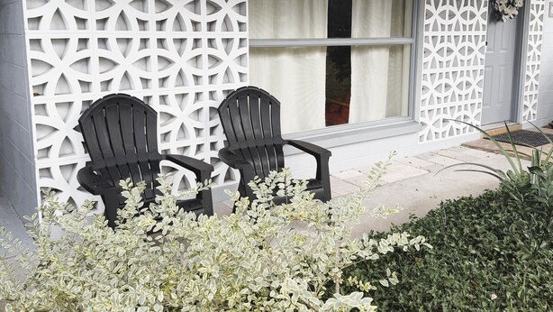 Ett hus med en vit, dekorativ skärm, två svarta Adirondack-stolar och ett fönster med vita gardiner.