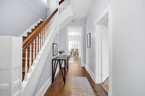 A hallway with a wooden staircase, a dining area in the background, and a rug on the floor.
