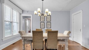 Une salle à manger avec une table blanche, quatre chaises en osier, un lustre et des œuvres d’art encadrées accrochées au mur.