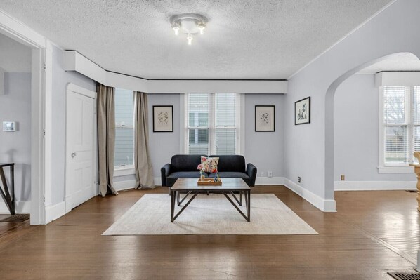 A spacious living room with hardwood flooring, a dark-colored sofa, a coffee table, and a large window with blinds.