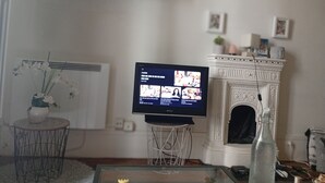 A living room with a television on a stand, a fireplace, a small table with a vase, and a shelf with books and decorative items. - Renting a room in a 2 room apartment (Paris)