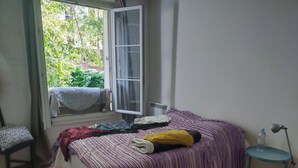 A bedroom with a bed, a chair, a small table, and a window with a view of greenery outside. - Renting a room in a 2 room apartment (Paris)