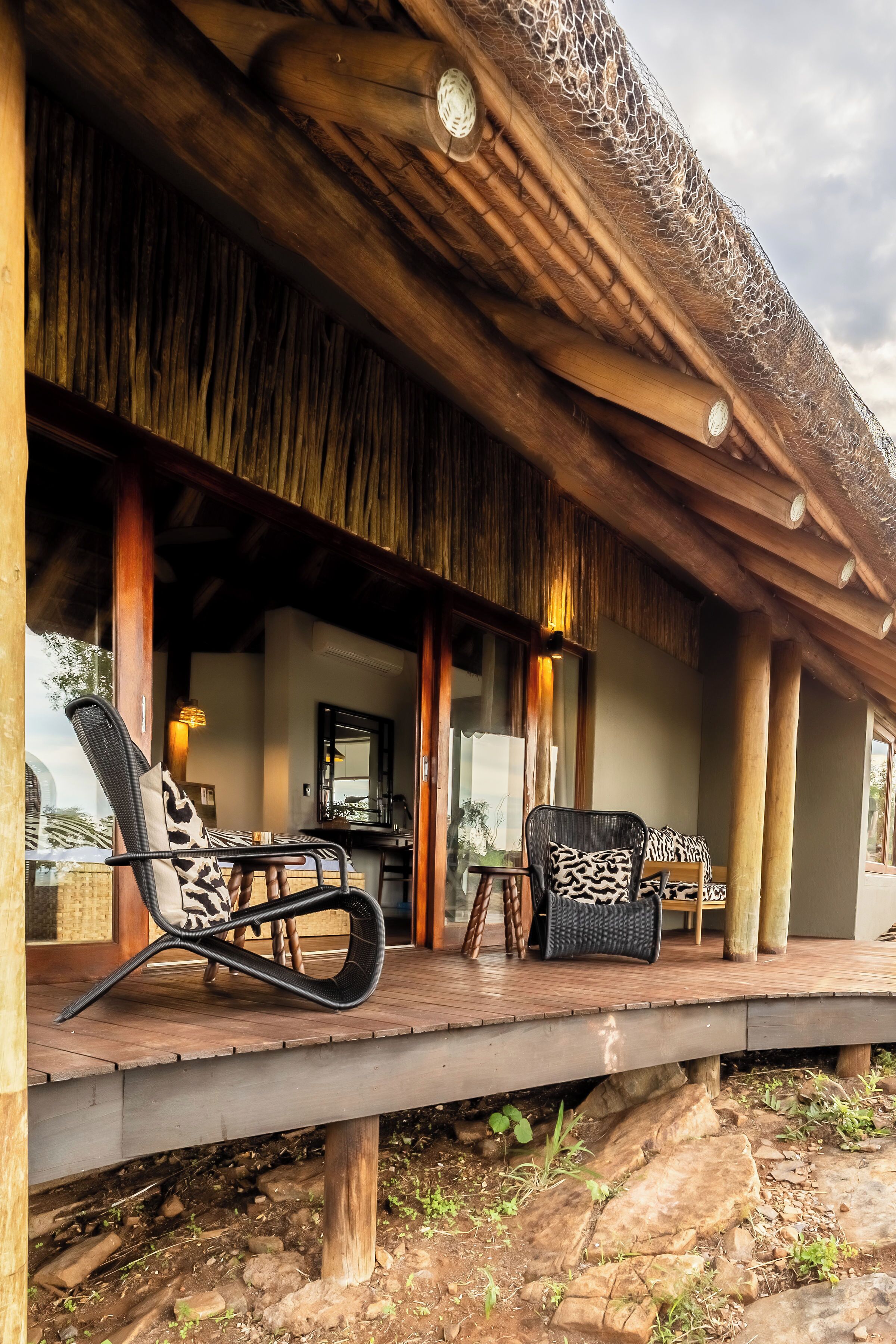 A wooden veranda with thatched roof, outdoor seating, and a view of the interior.