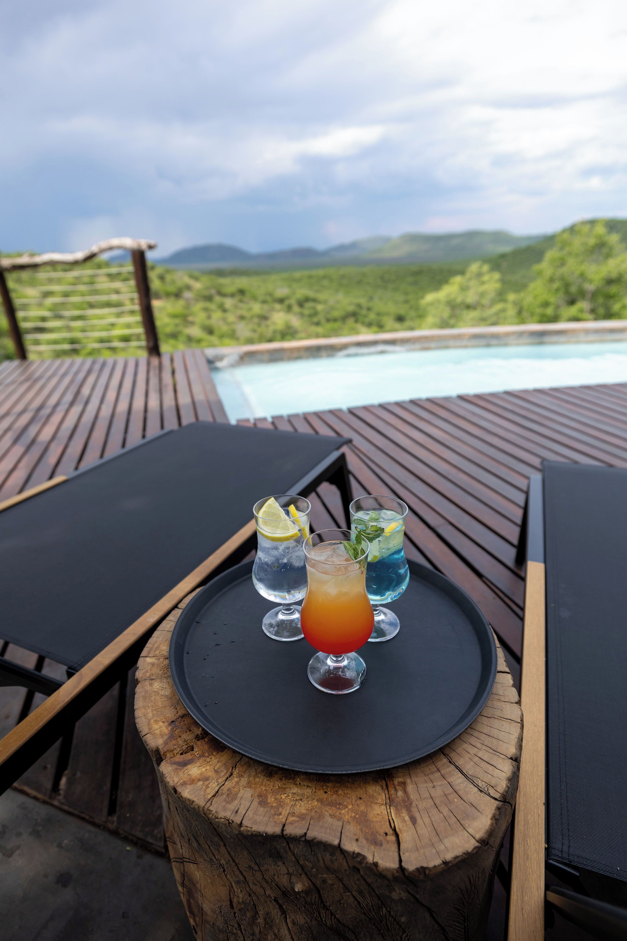 Une terrasse en bois avec une table ronde sur laquelle sont posées trois boissons, donnant sur une piscine et une végétation luxuriante.