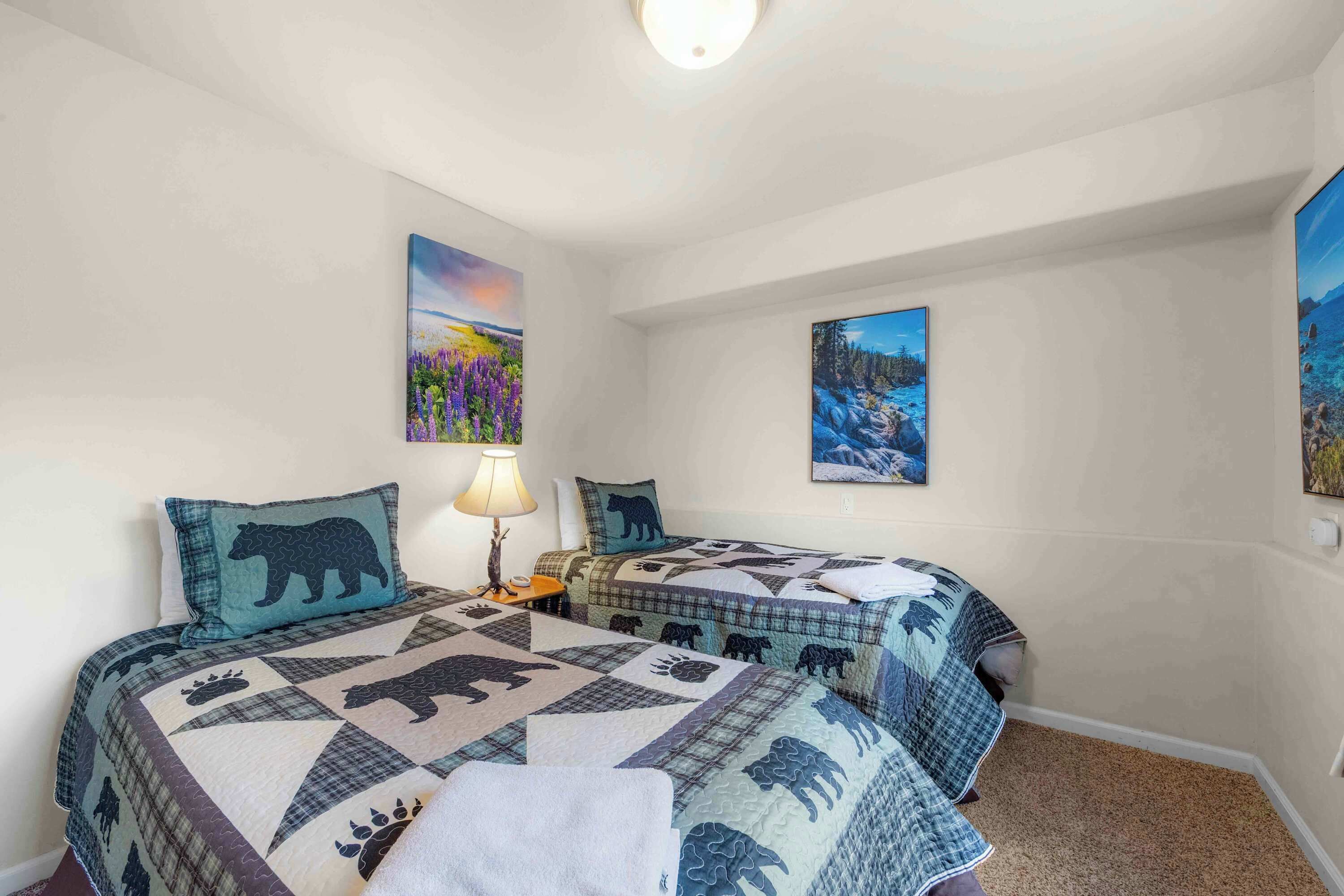 A bedroom with two beds, each with a bear-themed quilt, a nightstand with a lamp, and wall art depicting nature scenes.
