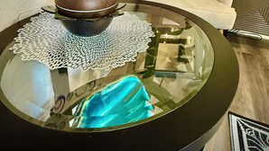 Una mesa de centro redonda de vidrio con un mantel decorativo, un florero marrón y un paño azul. Un sofá beige con un único cojín se ve al fondo.