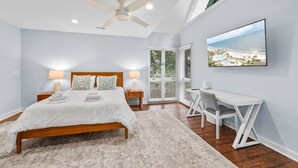 Un dormitorio con cama, mesitas de noche, escritorio, silla, ventilador de techo, ventana con persianas y televisor en la pared.