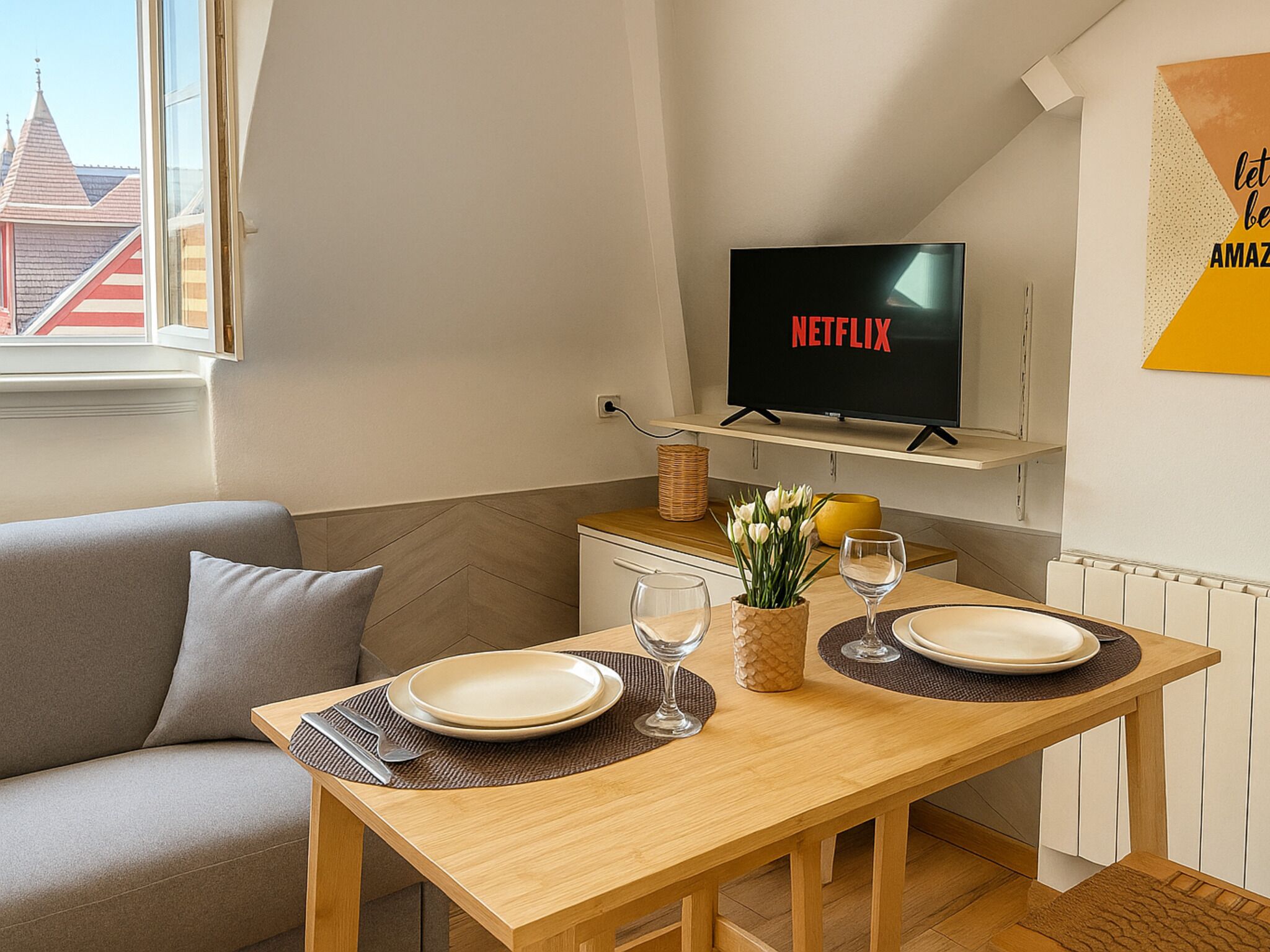 Un salon moderne avec une table à manger en bois, préparée pour deux personnes, un canapé, un téléviseur fixé au mur et une fenêtre donnant sur des immeubles.