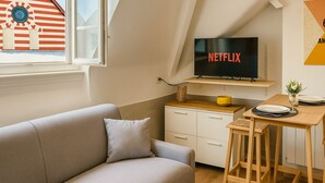 Een gezellige woonkamer met een bank, een aan de muur bevestigde televisie, een klein tafeltje en een raam met uitzicht op een gebouw.