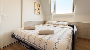 Een net opgemaakt bed met een bedsprei met patroon en twee kussens, een raam met gordijnen en een schilderij aan de muur.