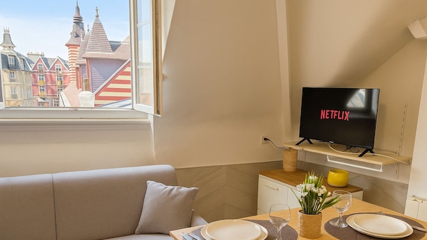 Een moderne woonkamer met een bank, een eettafel gedekt voor twee personen, een televisie op een standaard en een raam met uitzicht op kleurrijke gebouwen.