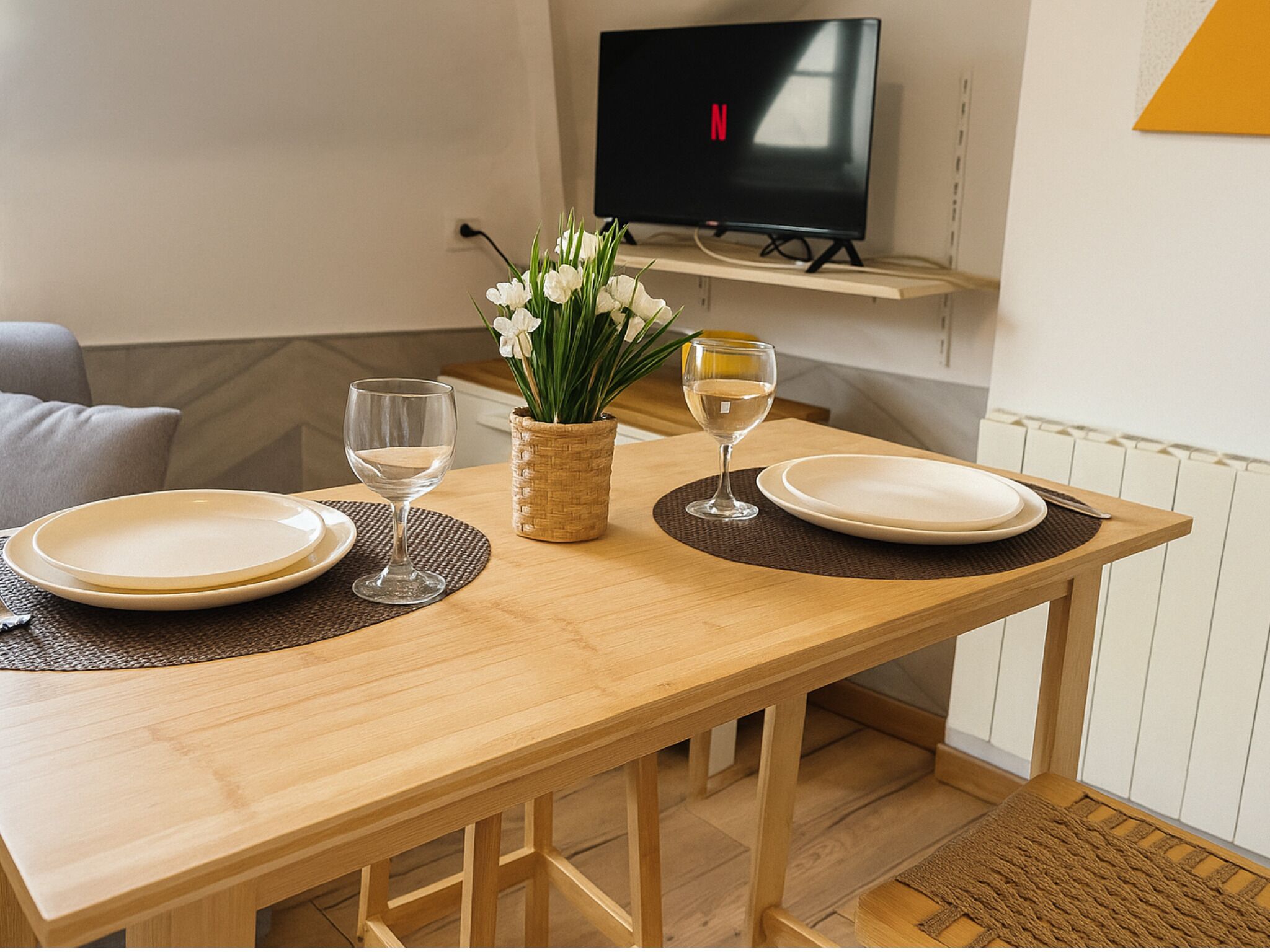 A dining table set with plates, glasses, and a vase of flowers in a room with a TV mounted on the wall.