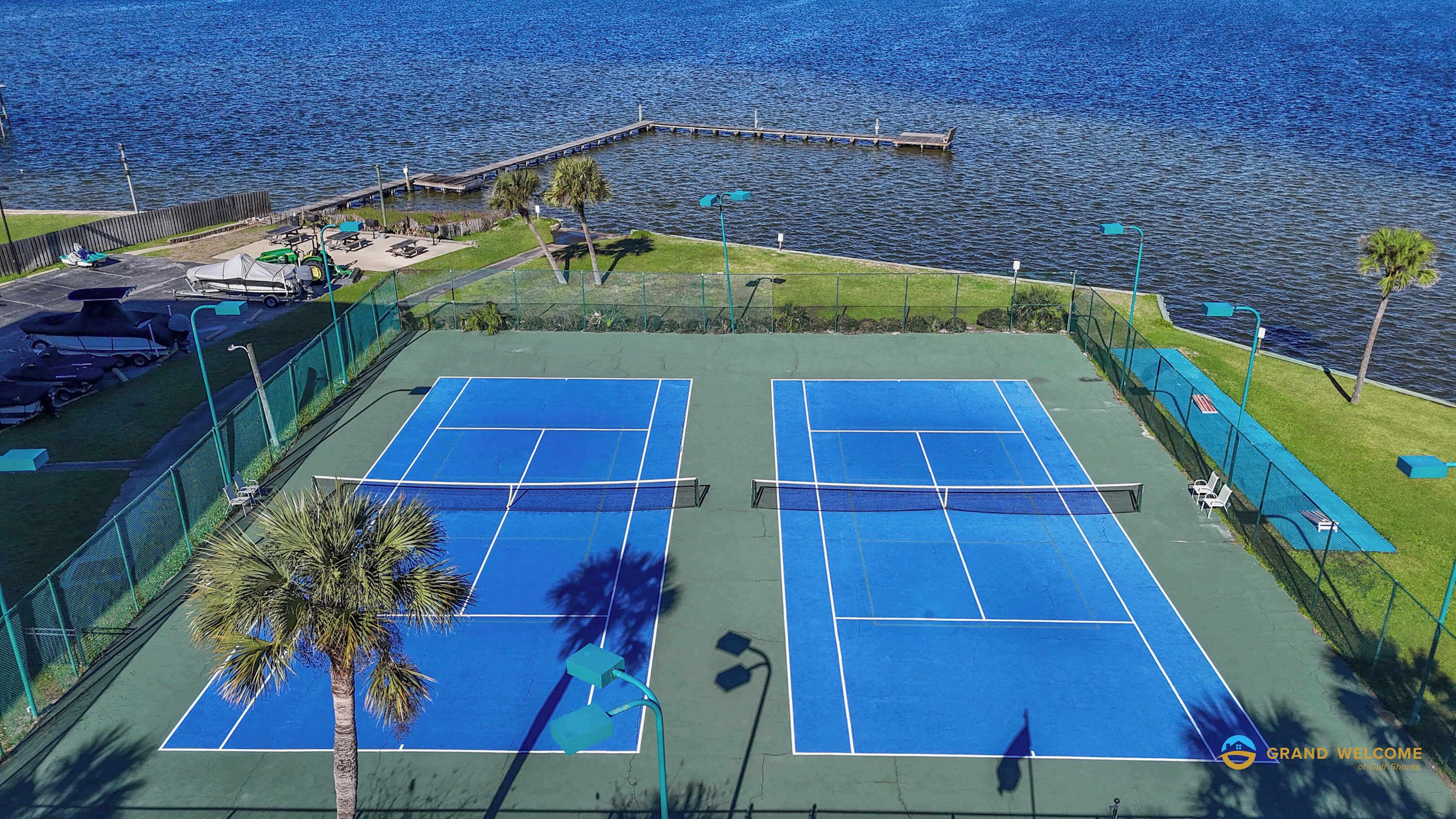 A tennis court with two courts, surrounded by a fence, with a dock and boats in the background.